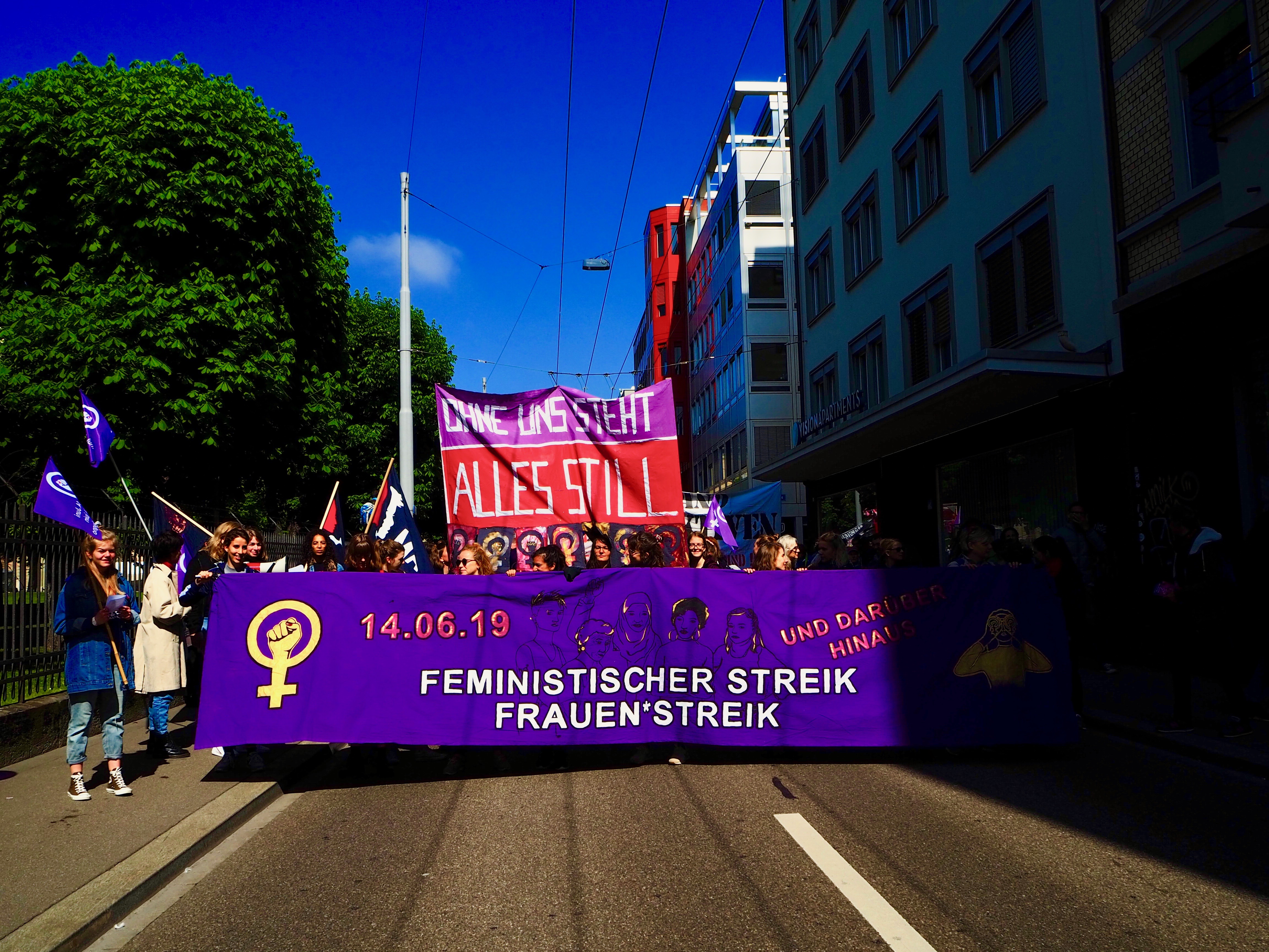 Foto vom Fronttransparent des Frauen*streikblocks. Darauf steht "Feministischer Strei / Frauen*streik 14.Juni 2019" Dahinter ein Transparent wo draufsteht "Ohne uns steht alles still".