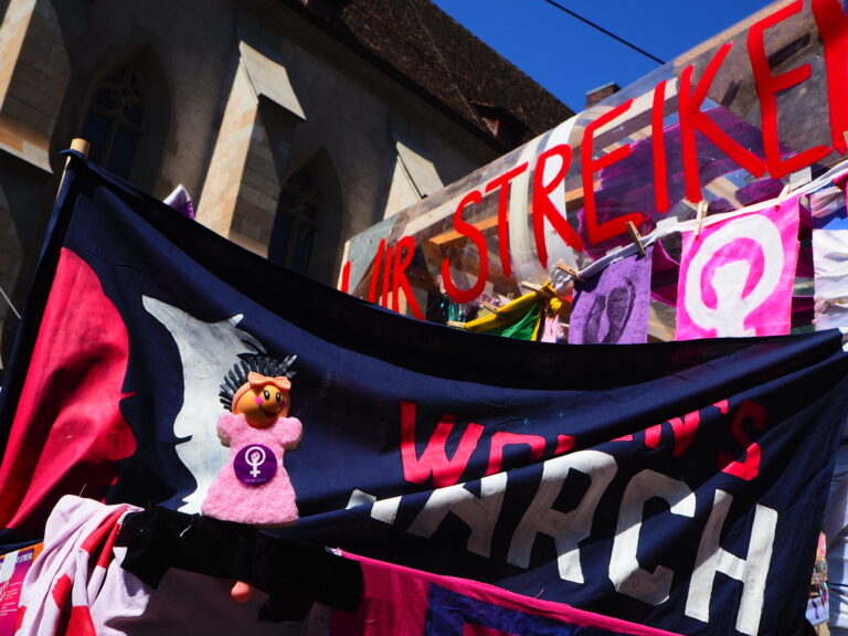 Foto vom Frauen*streikkblock am 1. Mai. Viele Transparente und zuvorderst Violetta, eine Geschirrspülbürste in Form einer Frau mit einem Schwammkleid, die befreut wurde und nun Teil des Streikkollektivs ist.