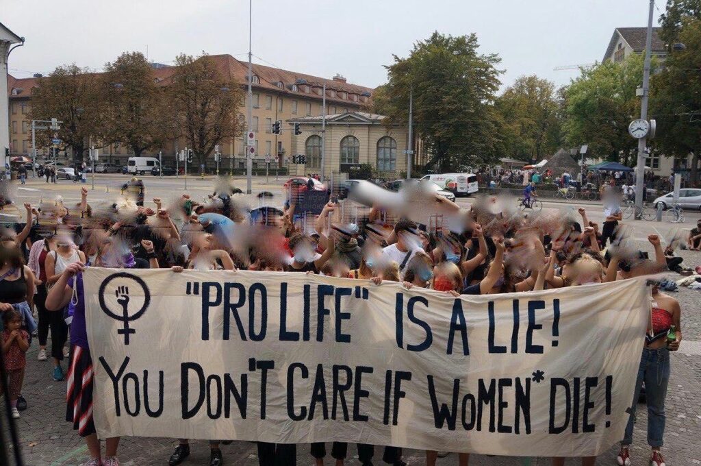Ein Solifoto auf dem Ni Una Menos Platz in Zürich, mit dem Transparent im Vordergrund auf dem steht "'Pro Live' is a lie! you don't care if women* die!"