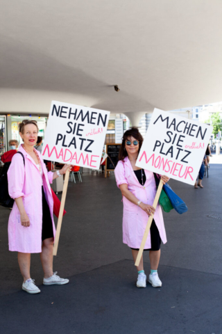 Zwei Personen in pinken Mänteln mit Schildern: "Nehmen Sie (endlich!) Platz Madame!" und "Machen Sie (endlich!) Platz Monsieur!".