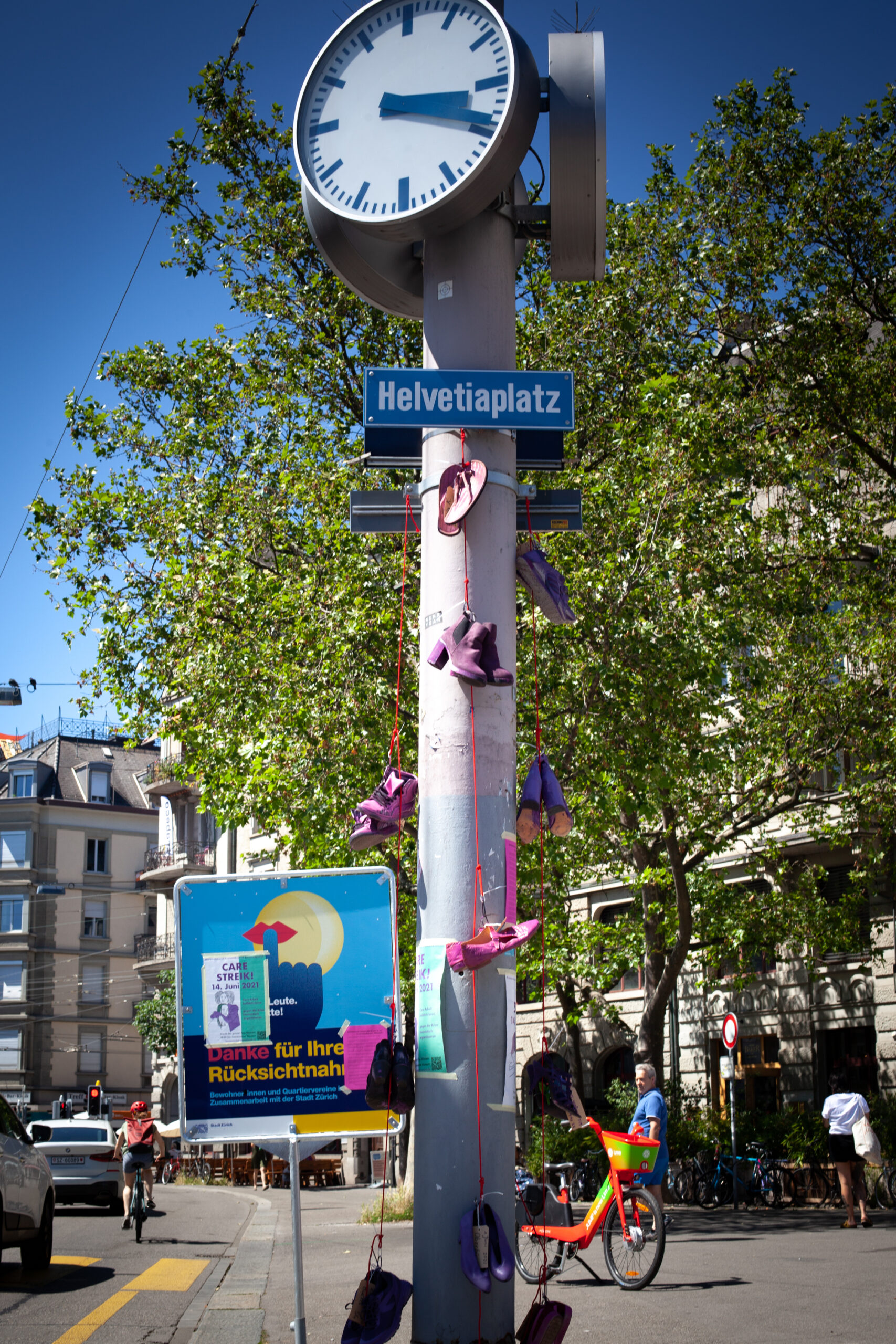 Die Uhr und Beschriftung des Helvetiaplatz mit allen 14 Schuhen, die in Memoria an ermordete FLINT*-Personen dieses Jahr in der Schweiz erinnert. Die Schuhe sind violett oder pink angemalt.
