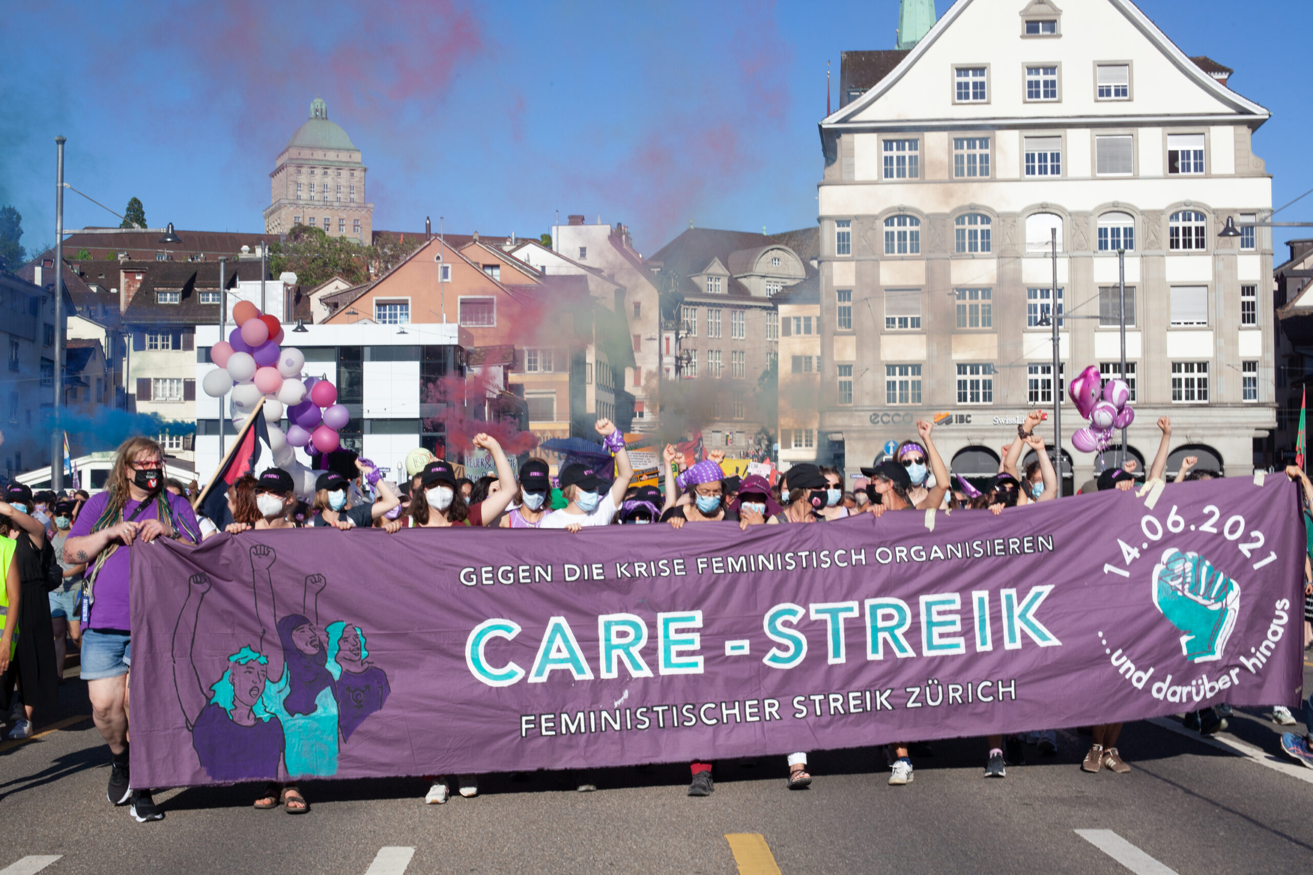 BIld der Demonstration auf der Rudolf-Brun-Brücke. Personen tragen das Frontbanner, auf dem steht: Gegen die Krise feministisch organisieren. CARE STREIK. Feministischer Streik Zürich. 14.06. 2021 ... und darüber hinaus.