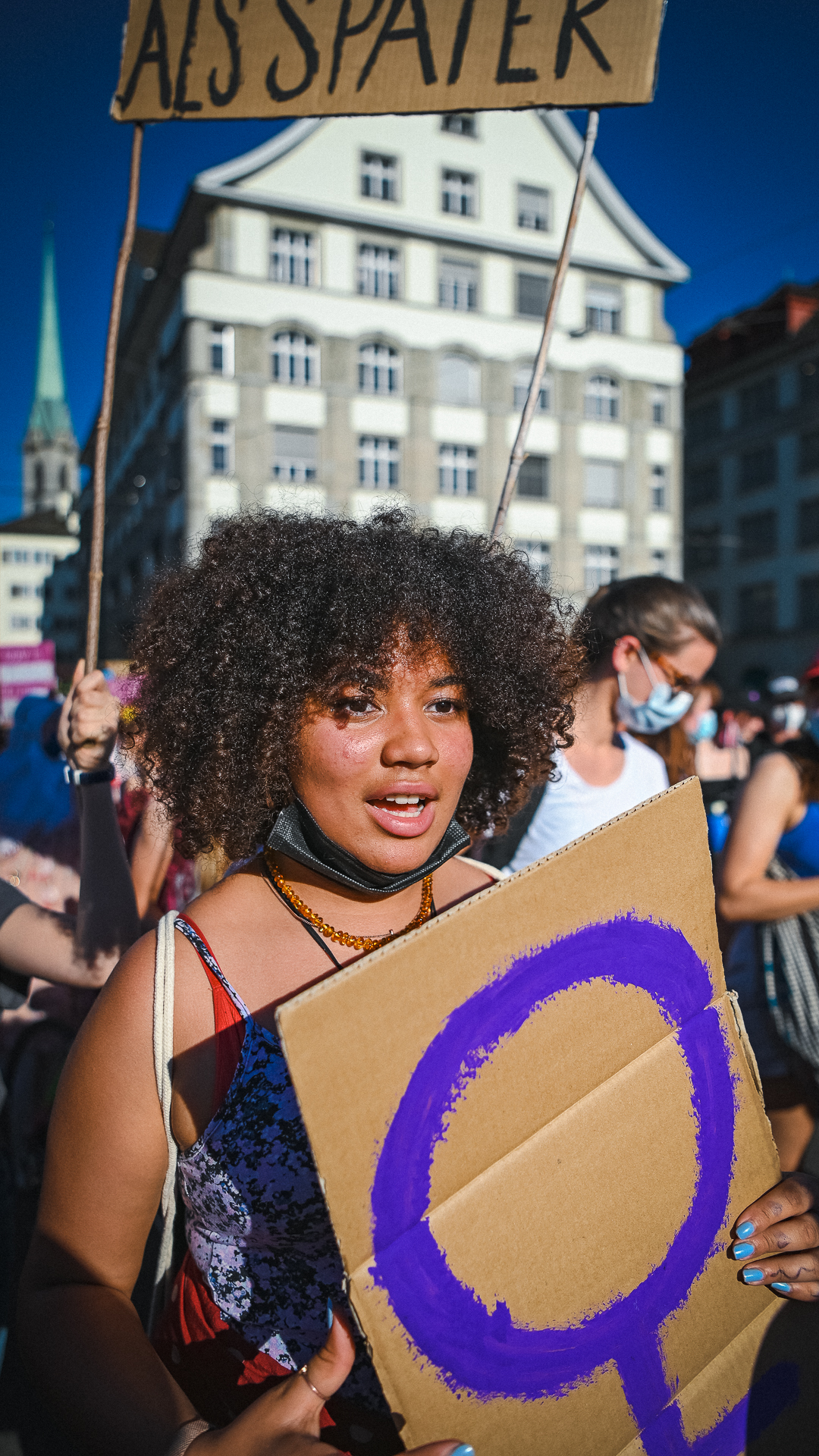 Ein Foto einer Frau mit dunklen Locken, die ein Schild mit dem violetten Venussymbol in den händen hält. Inmitten der Demo am 14. Juni 2021, sie ruft etwas