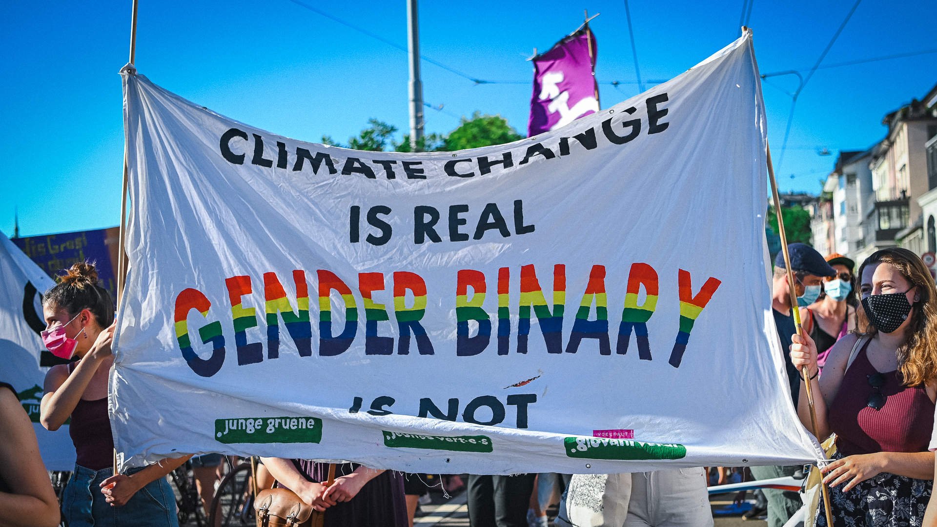 Ein Transparent in der Demo auf dem Limmatquai. Darauf steht: "Climat Change is real Gender Binary is not" und das logo der jungen grünen. "Gender Binary" ist in Regenbogenfarben geschrieben.