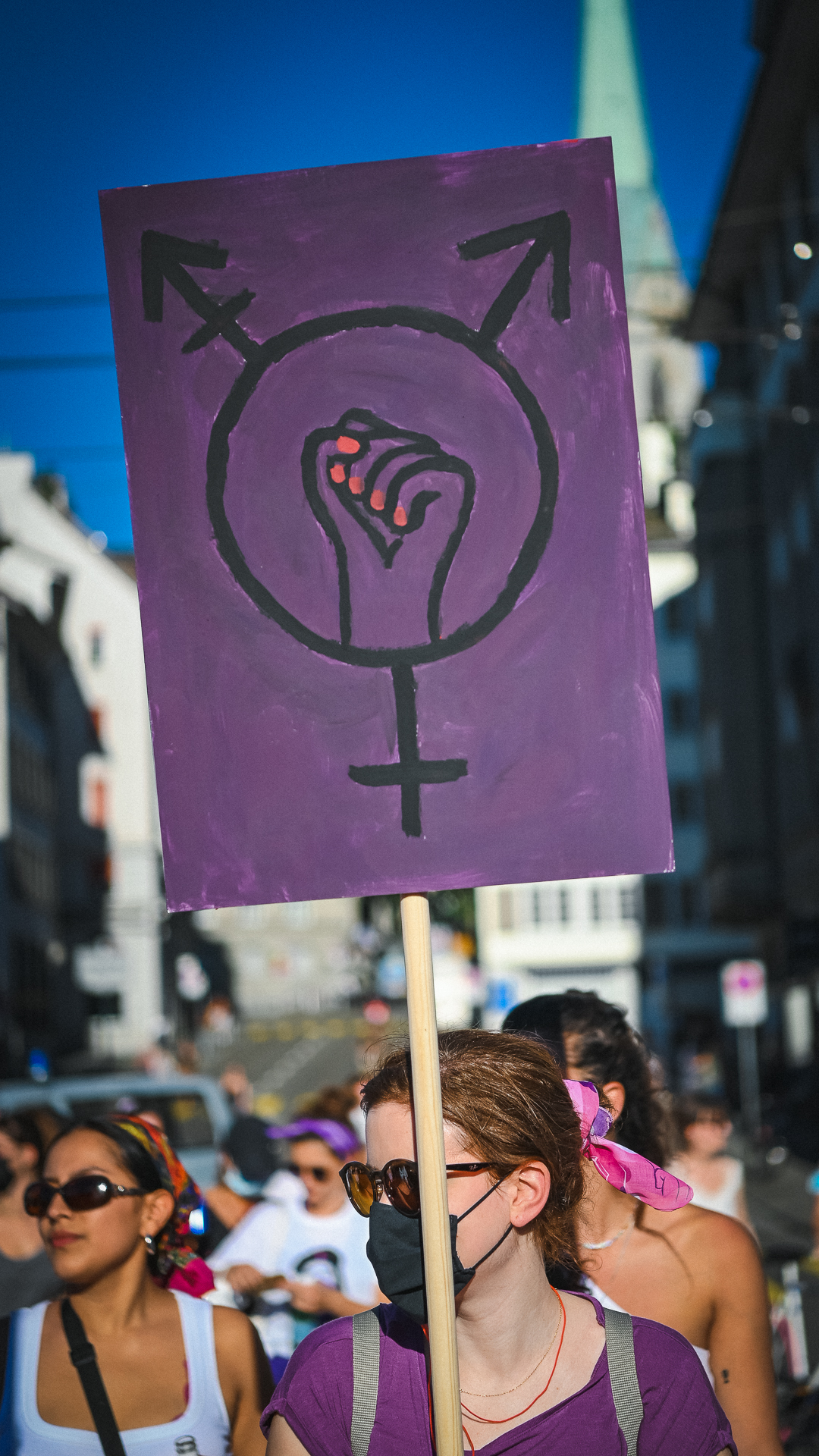 eine Person in der Demo in violettem Shirt hält hein violettes Pancarta hoch mit dem queerfeministischen Symbol mit einer Faust in der mitte mit rot lackierten Fingernägeln. Das Pancarta ist im Fokus