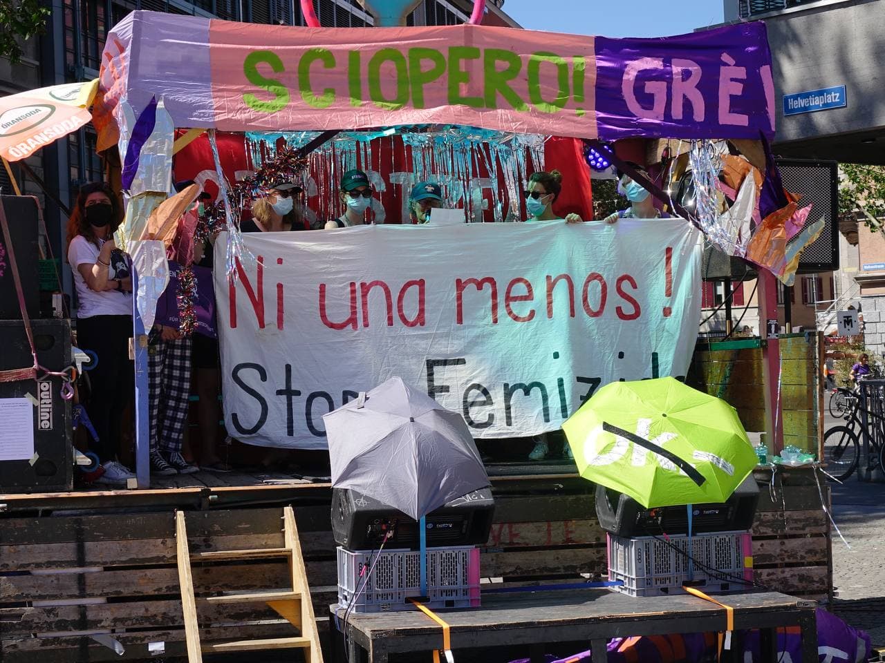 Foto der Bühne auf dem Ni UNa Menos Platz, ganz viele FLINTA mit Sonnebrille Caps und Maske halten ein Transparent hoch auf dem steht "Ni una menos! Stopp Femizide" eine Frau hält eine Rede. Die Bühne ist mit Lametta und Banner verziert auf dem Steht "Sciopero! Grève! …"