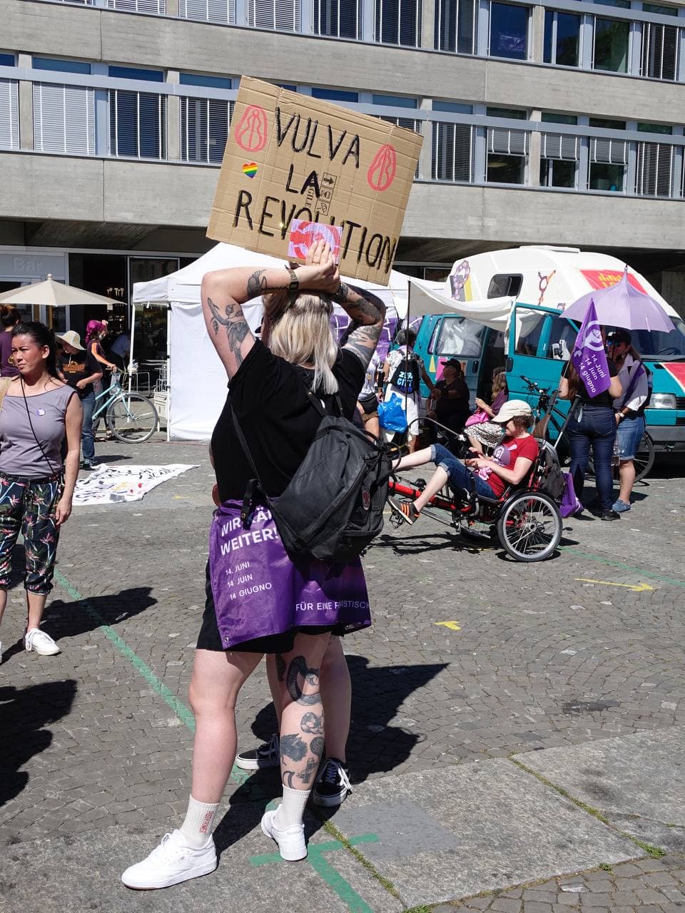 Eine Person hätl auf dem Ni Una Menos Platz ein Schild hoch auf dem Steht "Vulva La Revolution" verzierz mit stilisierten Zeichnungen von Vulvas. Sie hat eine Streikfahne umgebunden. Im Hintergrund ist der Wagen von Radio Lora zu sehen.