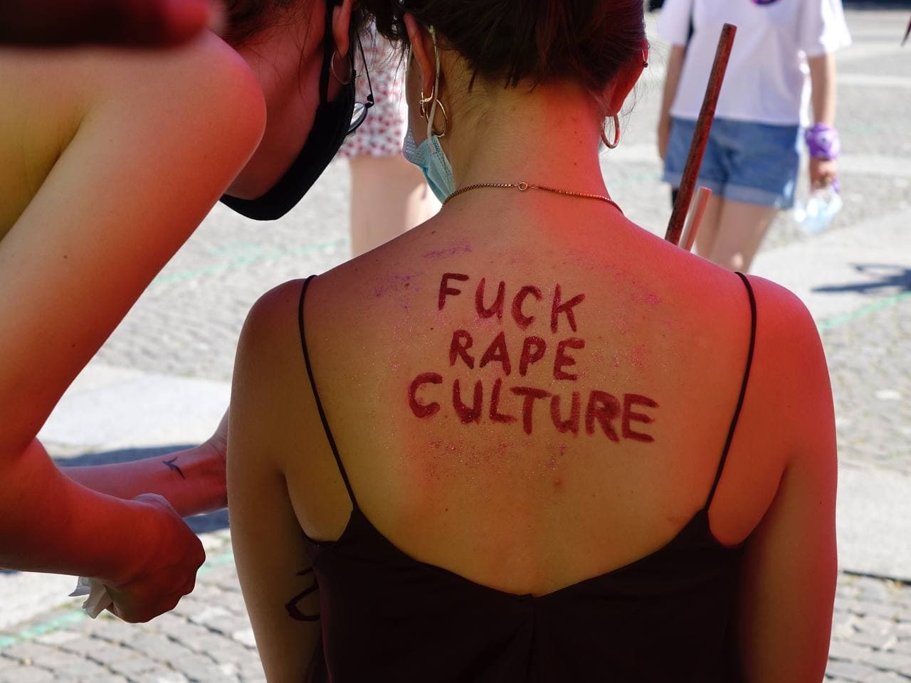 Foto des Oberkörpers einer Person von hinten, welche ein Träger-Shirt trägt. auf ihrem Freien Rücken steht mit rot und glitzer "Fuck rape culture" eine weitere Person bückt sich zu ihr herunter und zeigt ihr was.