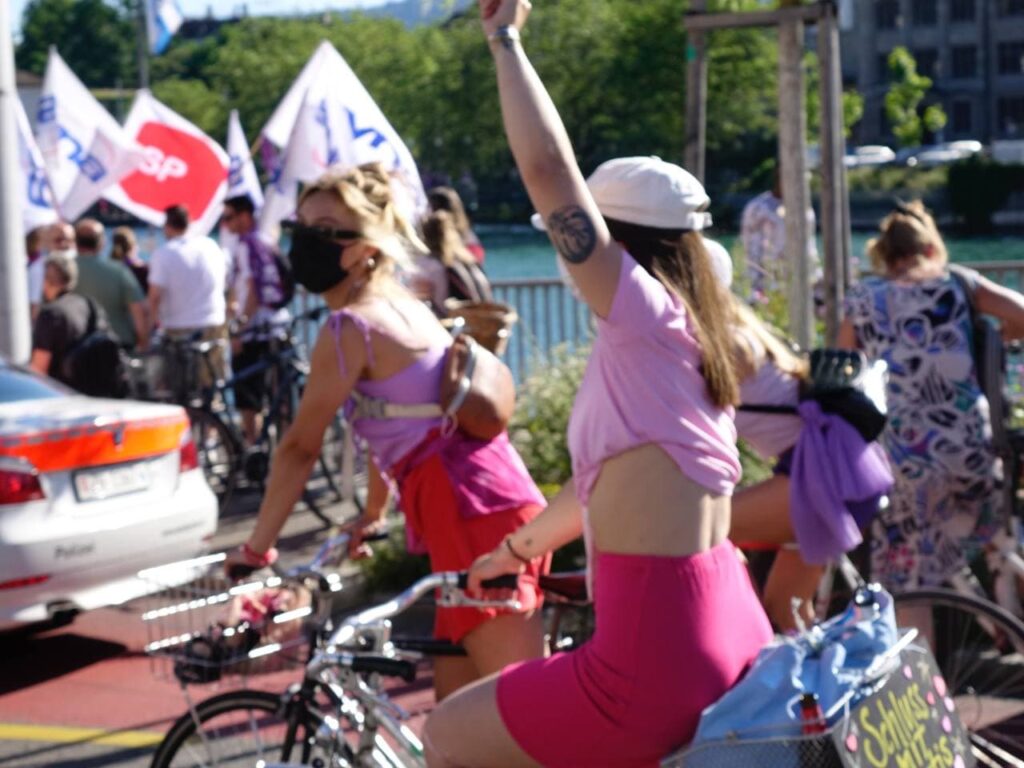 eine verschwommene Aufnahme von zwei Personen in Pink/roten Shorts und rosa Top auf dem Fahrrad, die vordere Person reckt die Faust in die Höhe. Sie fahren auf dm limmatquai, im Hintergrund ist eine Gruppe mit verschiedenenen Partei und Gewerkschaftsfahenen zu sehen