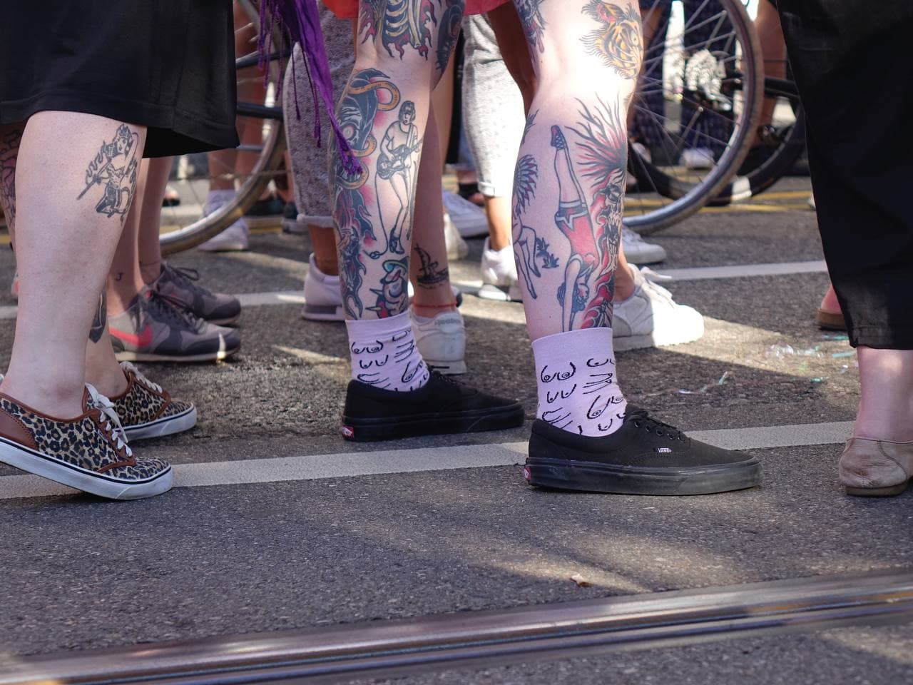 Nahaufnahme von vielen Beinen, zuvorderst zwei bunt tättowierte Beine die in schwarzen Schuhen stecken und rosa Sockn tragen auf denene viele Brüste aufzeichnet sind
