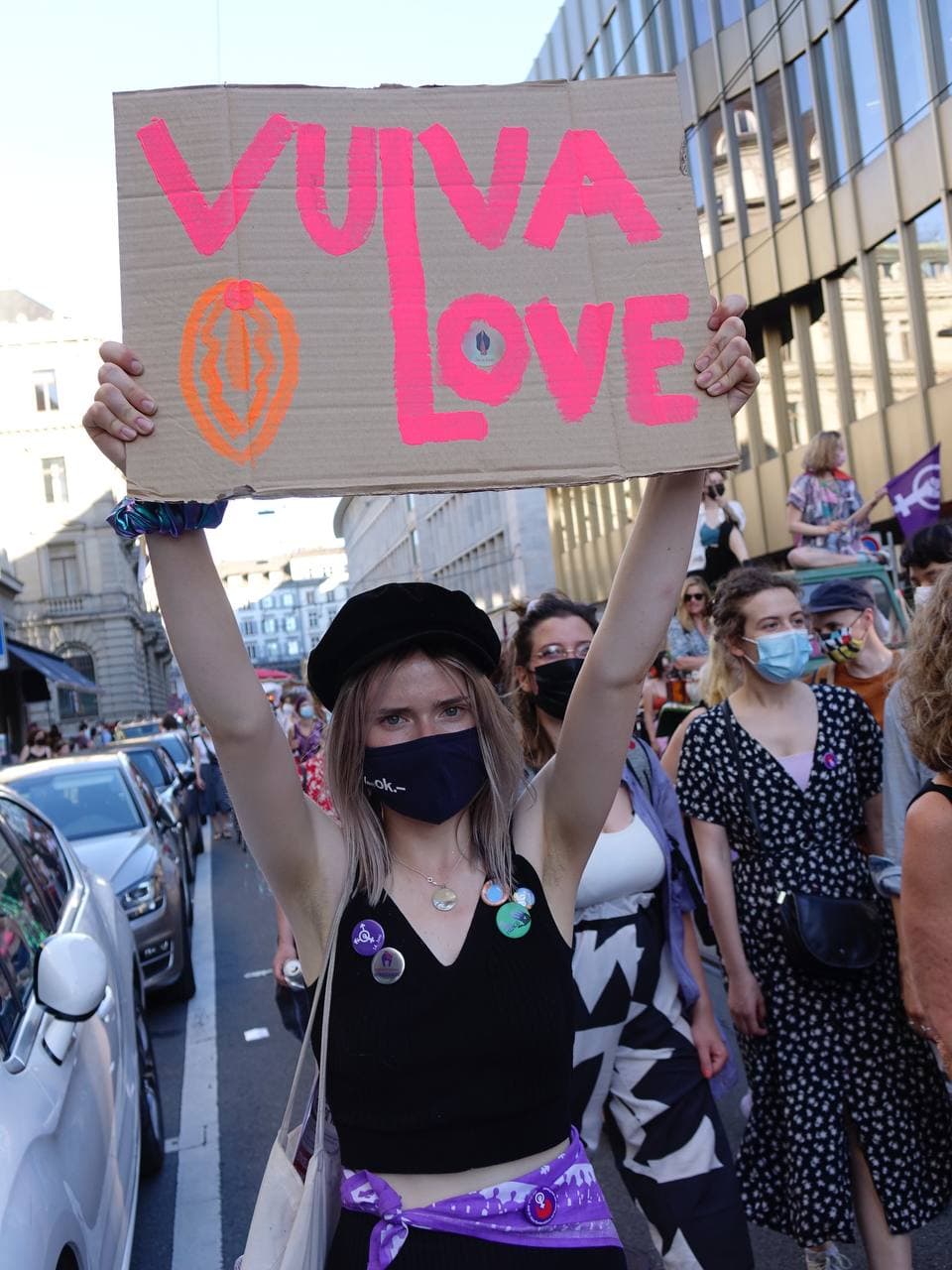 Eine Frau mit schwarzer Mütze und Schwarzer Maske und vieler feministischer Pins hält im Demozug stehend ein Schild hoch auf dem in Orange ein Vulva gezeichent ist und in Pink steht "Vulva Love" wobei das L von Vulva auch das L von Love ist. Die Person schaut direkt in die Kamera