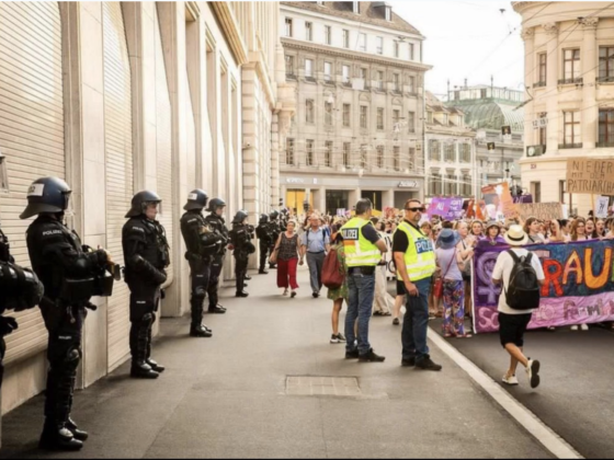 Ein Foto aus einer Gasse in Basel, rechts sehen wir den anmarschierenden feministischen Demozug mit einem Bunten Fronttranspi. Links ist dem Gebäude entlang die Polizie in vollmontur und z.T. mit Schrotgewehr bereit aufgereit.
